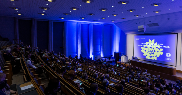 Yleiskuva vuosipäivän symposiumista. Auditorio, ihmisiä istumassa ja siniset valot.