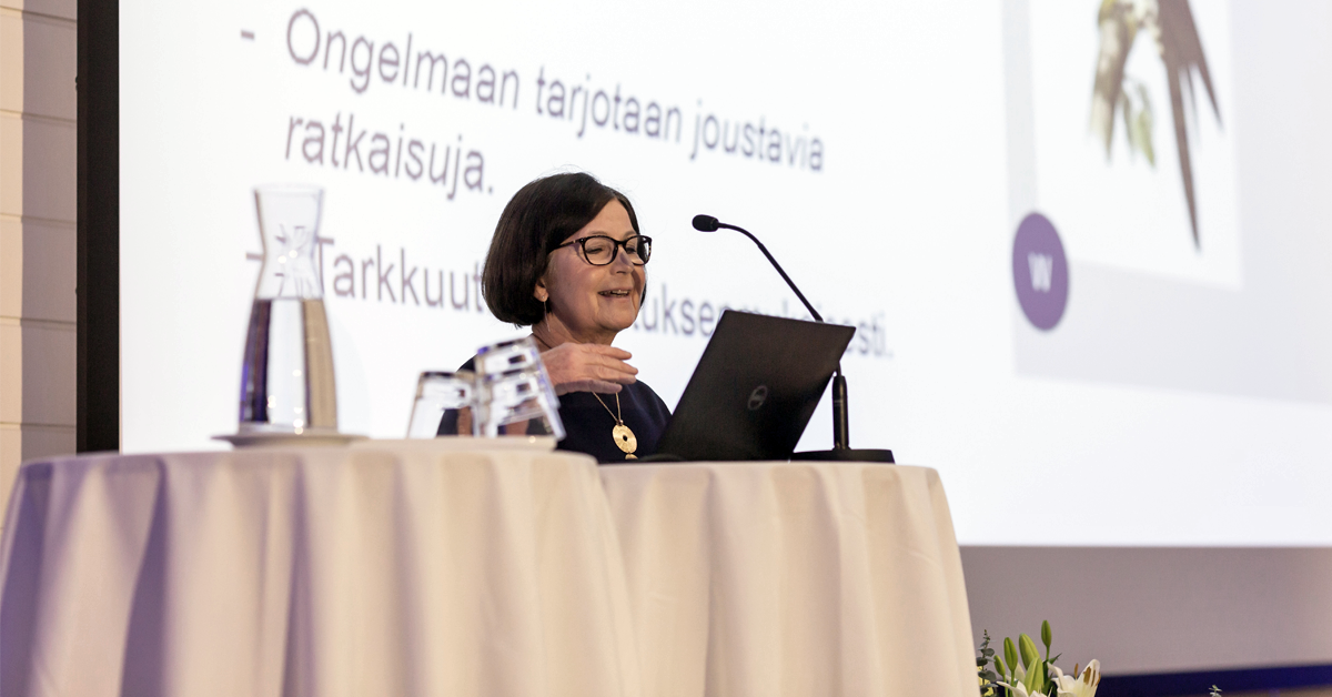 Kaisa Häkkinen lavalla pitämässä luentoaan, valkoiset pöydät ja tietokone, taustalla slidet.