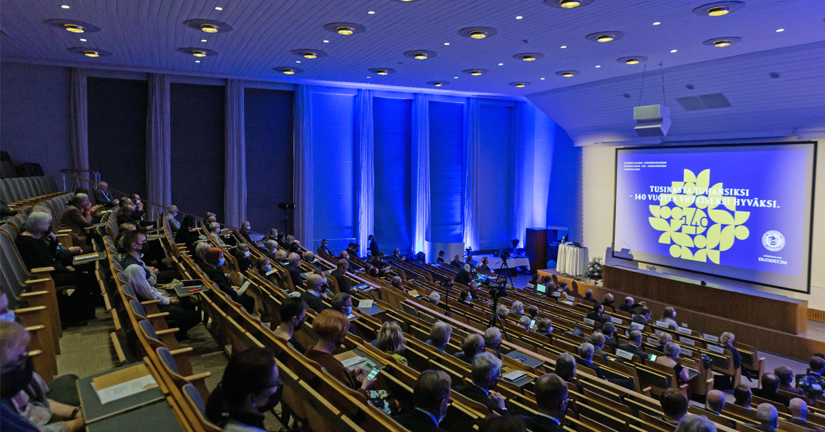 Kuva Ravintola Töölön auditoriosta, jossa ihmisiä katsomossa, sininen valaistus ja lavalle heijastettuna kultainen 140. vuosipäivän ruusuke.
