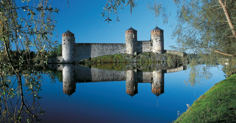 Olavinlinna auringonpaisteessa, heijastuu järven pinnasta. Terveyttä taiteesta -kulttuuriseminaarin 2023 markkinointikuva.