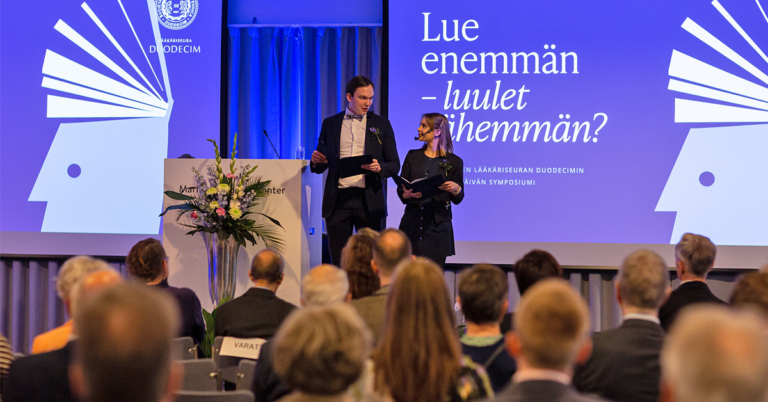 Duodecimin 141. vuosipäivän symposiumin yleisö etualalla ja juontajat taka-alalla lavalla.