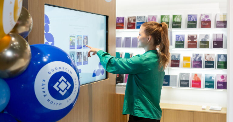 Nuori lääkäri pelaa Duodecimin osastolla juhlavuosien peliä. Edessä Käypä hoito 30 vuotta -ilmapallo ja taustalla Kustannus Oy Duodecimin kirjoja hyllyissä.
